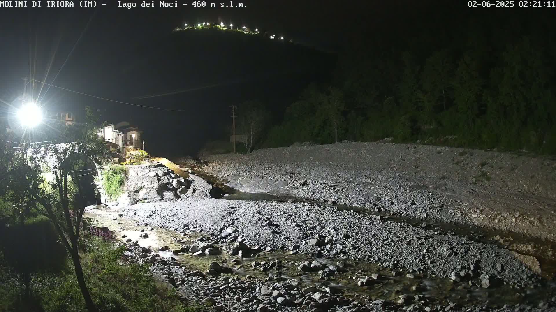 A rocky, shallow stream flows through a dark, nighttime landscape under artificial light, past a village nestled at the base of a hill.