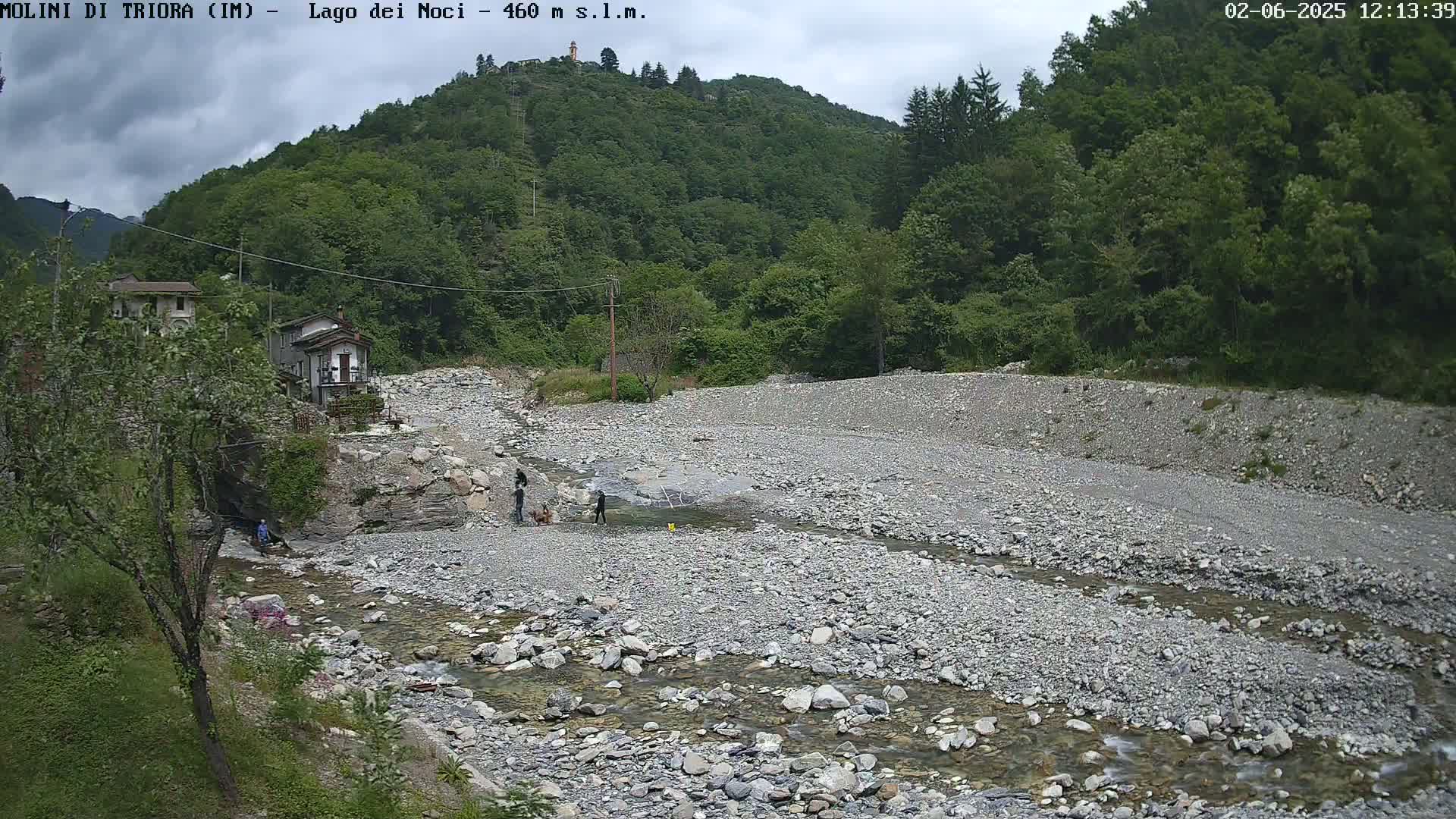 A mostly cloudy day reveals a rocky streambed with a slow-moving stream, bordered by lush green hills and a few small buildings.