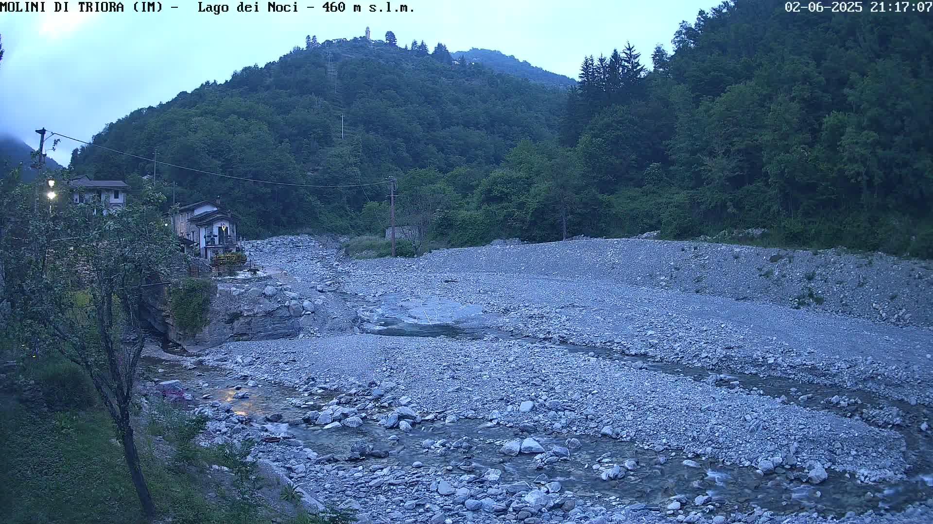 A rocky, shallow stream flows through a sparsely vegetated valley under a twilight sky, with a small village nestled at the edge of the valley.