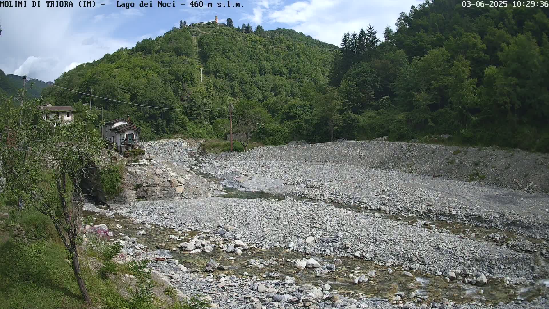 A shallow, rocky stream flows through a mostly dry riverbed beneath a partly cloudy sky, past a few buildings nestled at the base of a green hillside.