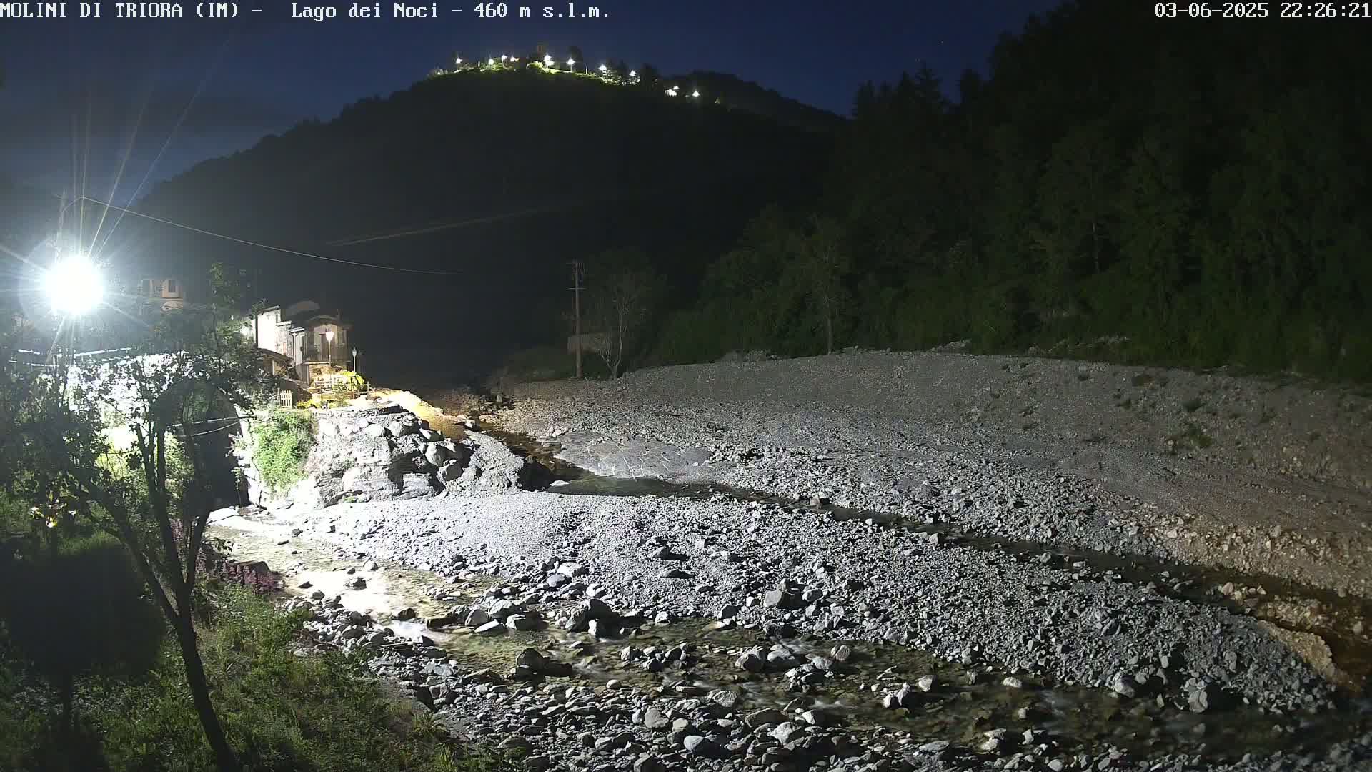 A rocky, shallow stream flows past a village at night, under a dark sky, with a hilltop illuminated in the distance.