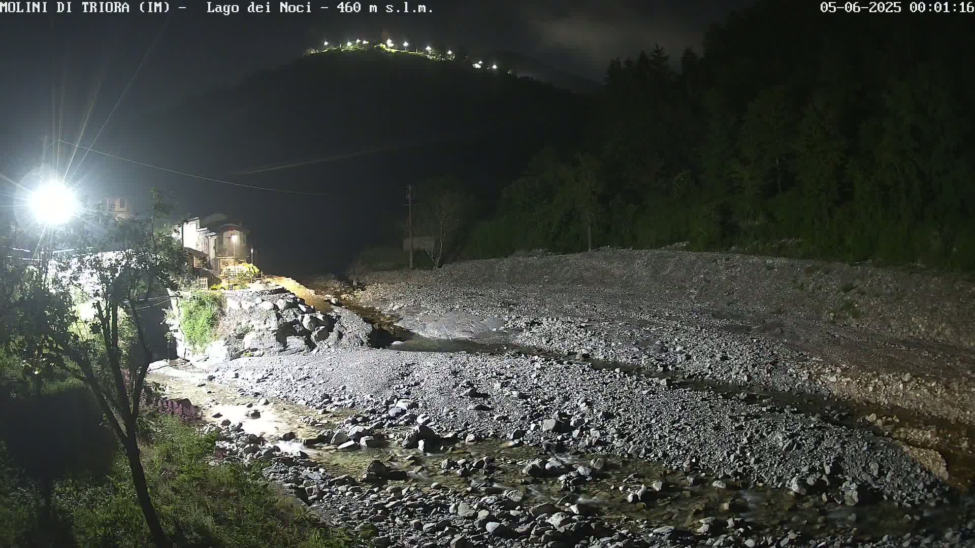 A rocky stream flows through a dark, nighttime landscape under artificial light, past a small village nestled at the foot of a hill.