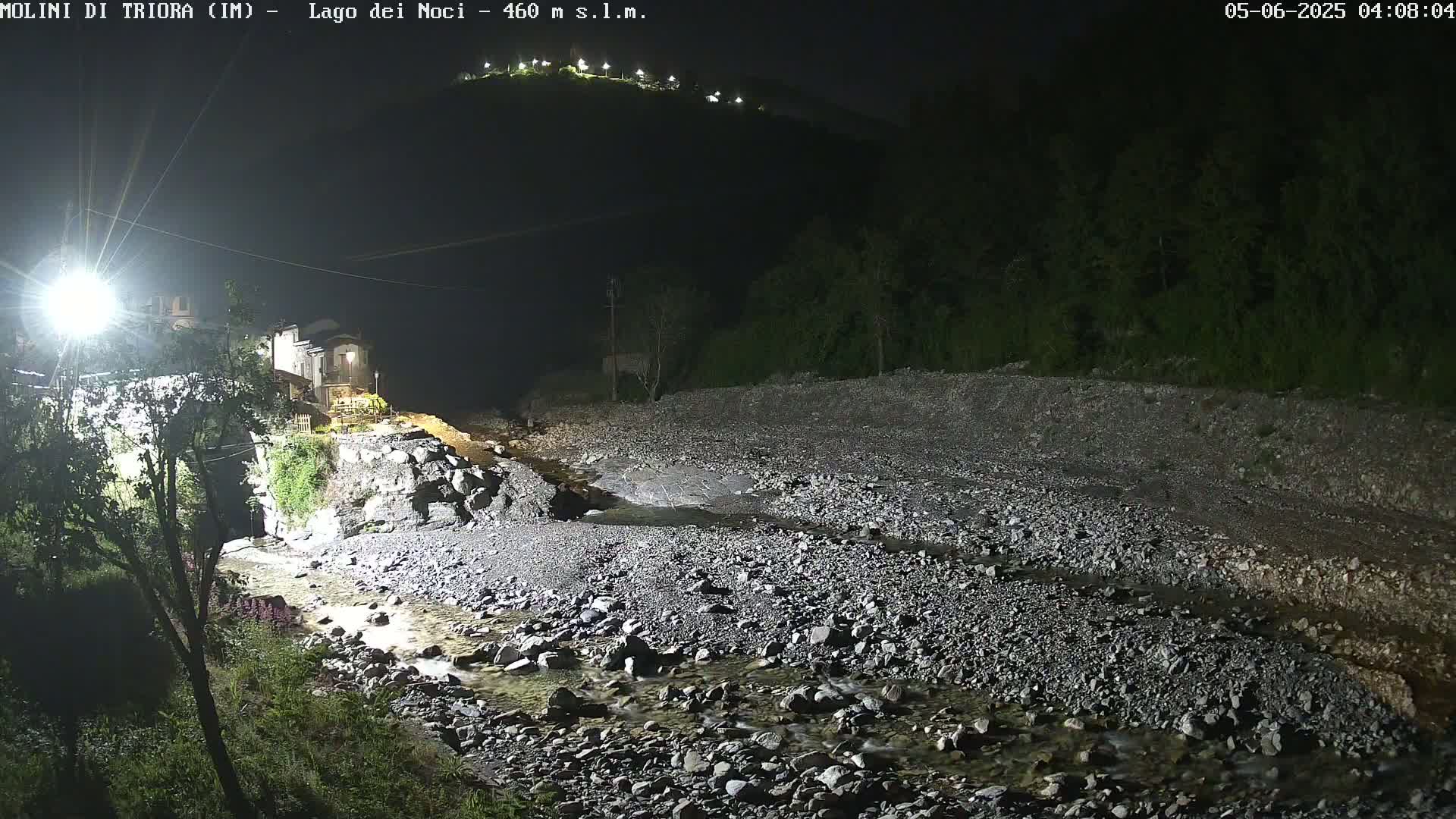 A rocky, shallow stream flows through a dark, nighttime landscape, past a small village illuminated by artificial lights on a hill.