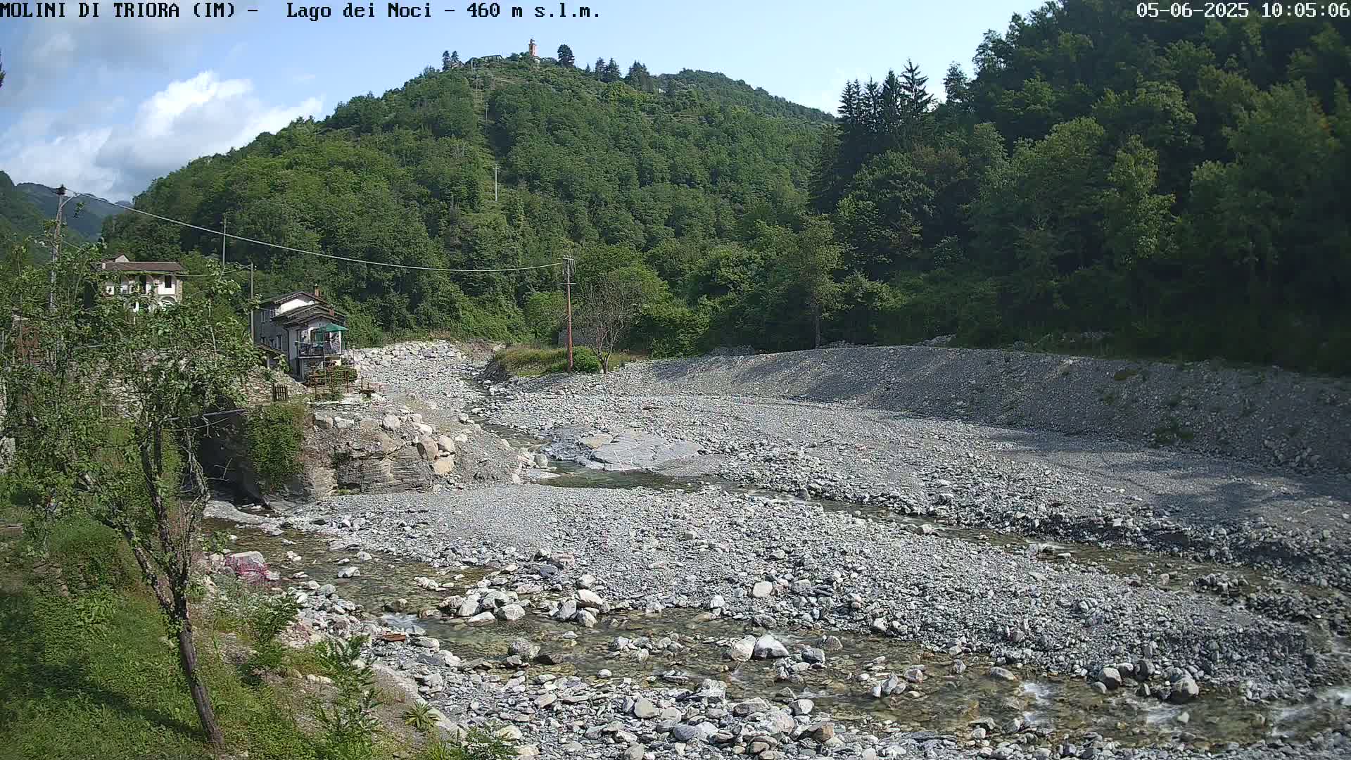 A rocky, shallow river flows through a dry riverbed, surrounded by lush green hills under a mostly clear sky.
