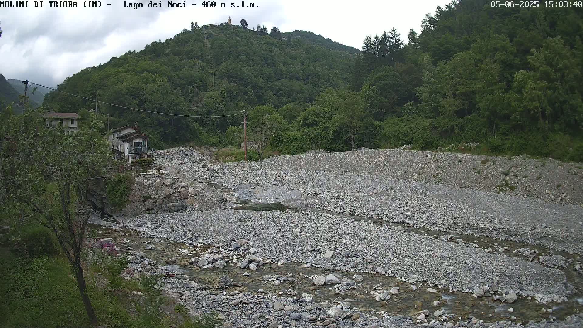 A shallow, rocky river flows through a dry riverbed beneath a partly cloudy sky, past a hillside covered in lush green vegetation and a few buildings.