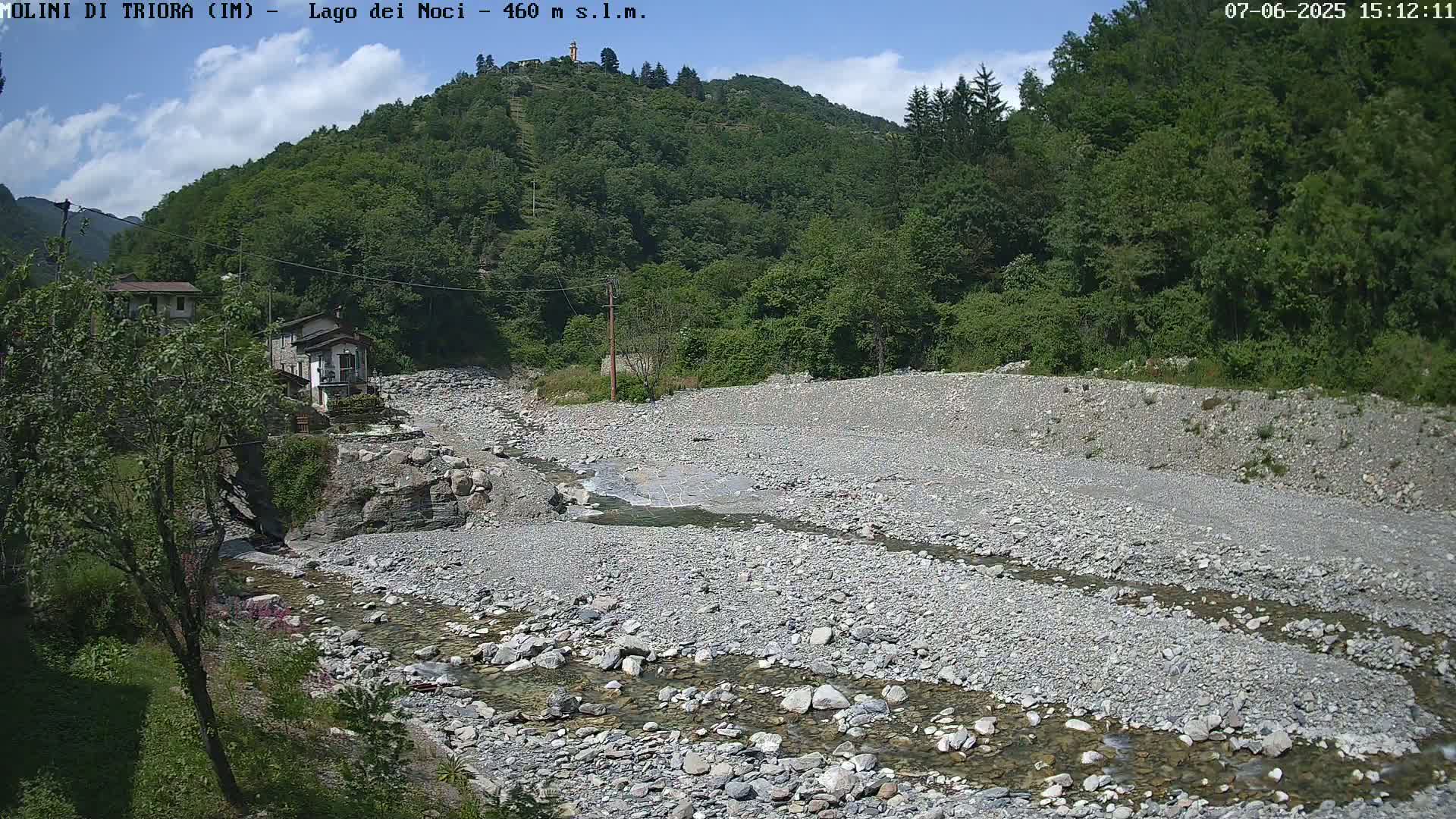 A shallow, rocky river flows through a dry, stony riverbed, surrounded by lush green hills under a partly sunny sky.