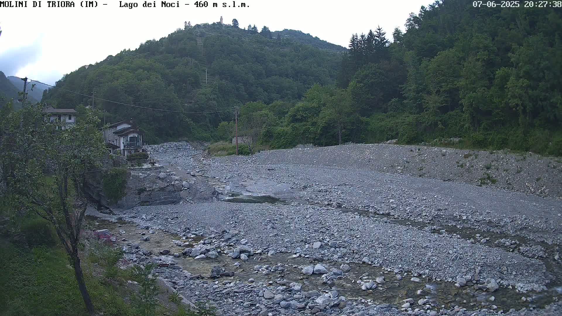 A rocky, shallow river flows through a valley under an overcast sky, past a few buildings nestled beside a lush green hillside.