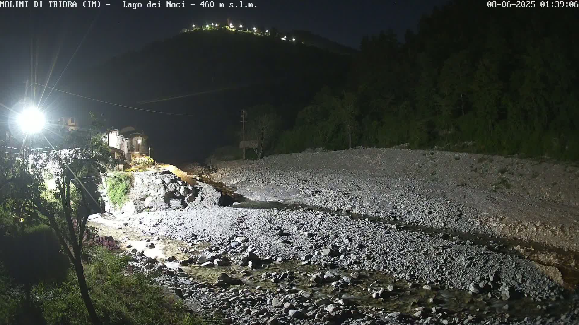 A rocky, shallow stream flows through a nighttime landscape under a clear, dark sky, past a building illuminated by artificial light, with a hill visible in the background.