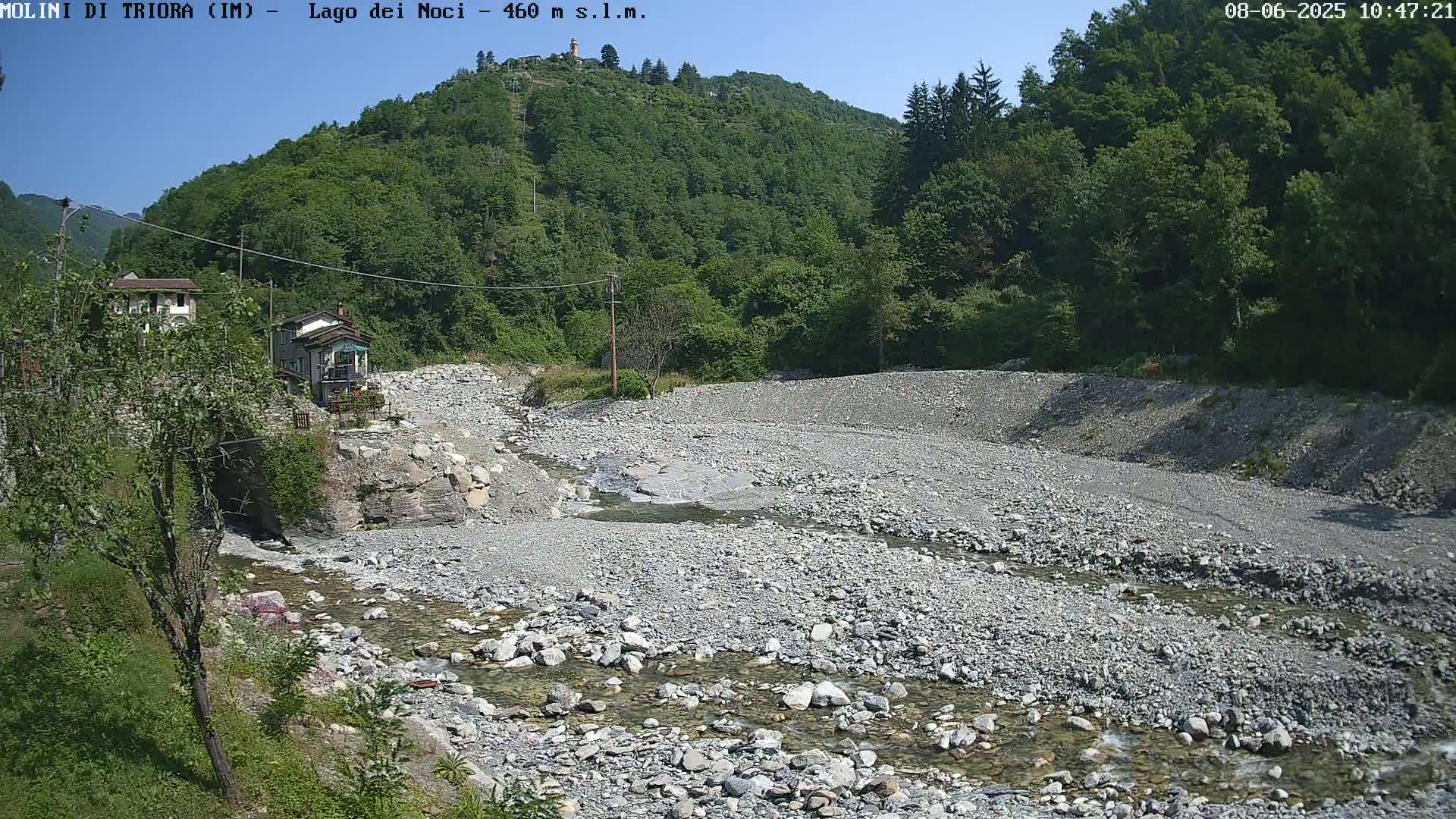 A rocky riverbed with a shallow stream flows through it, surrounded by lush green hills under a clear, sunny sky.