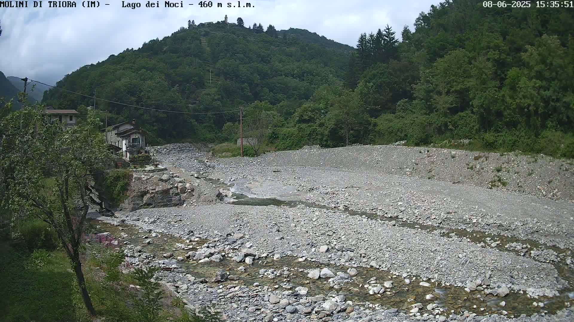 A rocky, shallow river flows through a mostly dry riverbed, past a small village nestled at the base of a lush green mountain under a partly cloudy sky.