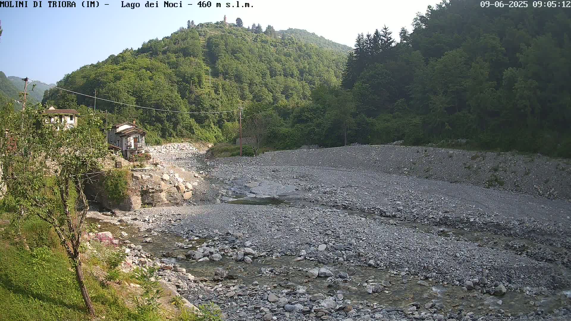 A mostly dry, rocky riverbed with a small stream flows through it, surrounded by green hillsides under a mostly clear sky.