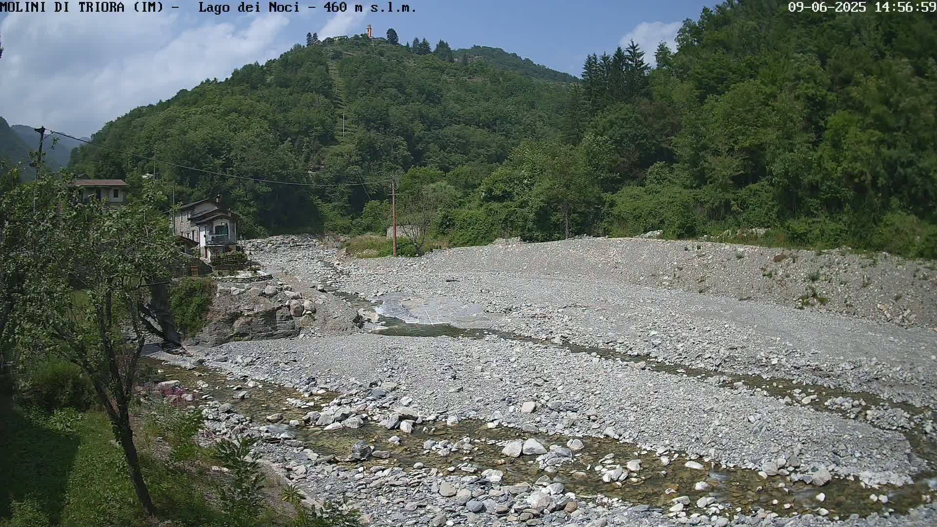 A shallow, rocky river flows through a dry, gravelly riverbed, nestled within a verdant, mountainous landscape under a mostly sunny sky.