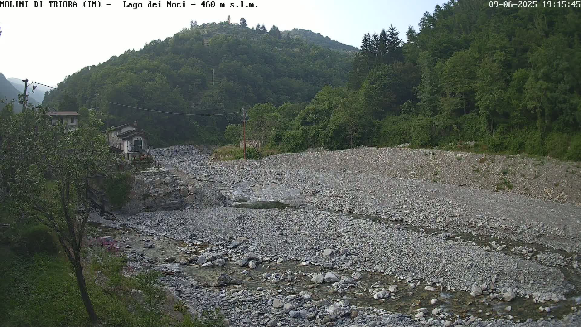 A rocky riverbed with a shallow stream flows through it, nestled between lush green hillsides under a mostly clear sky.