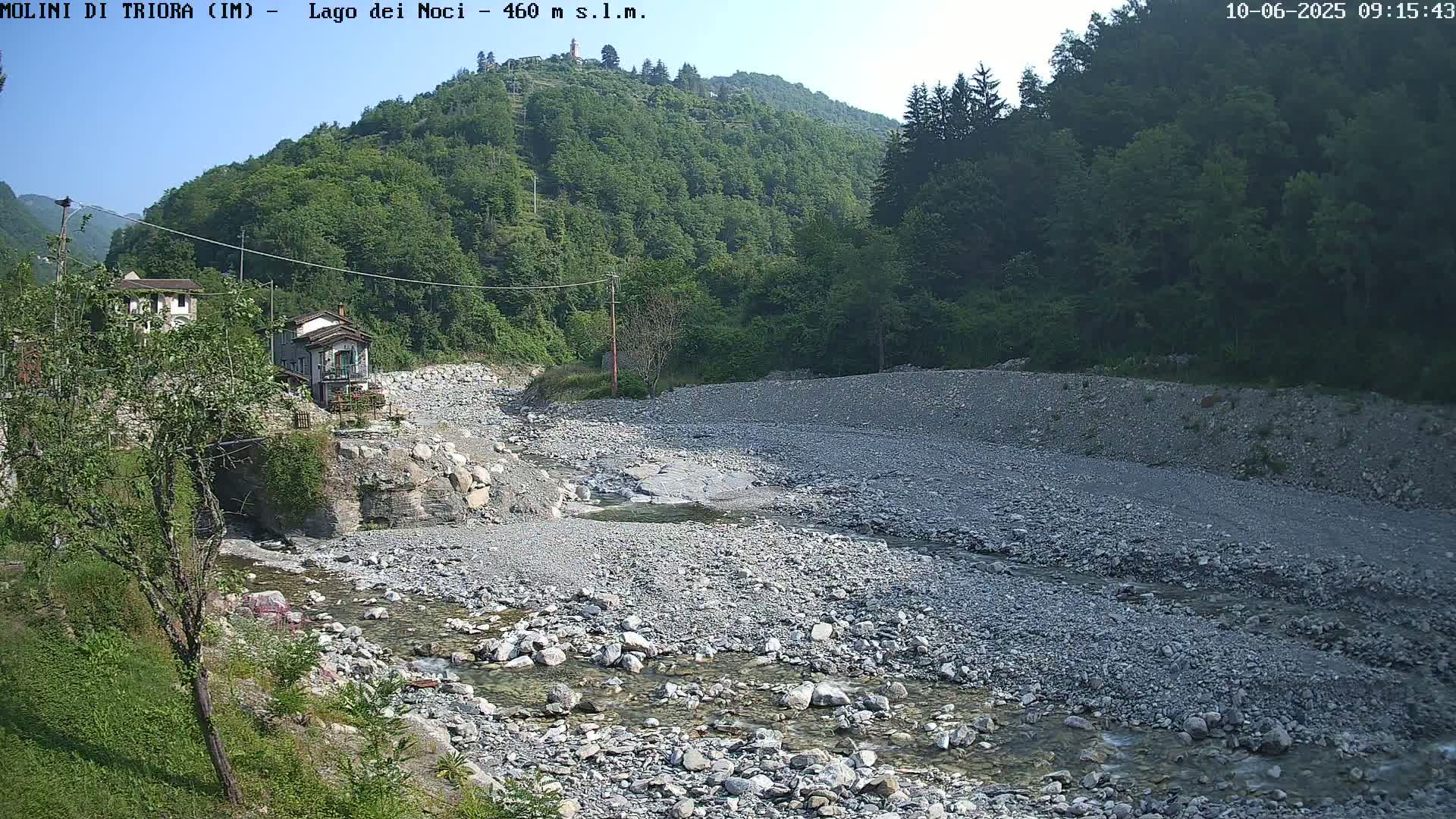 A shallow, rocky stream flows through a dry riverbed, past a small village nestled at the base of a lush green mountain under a clear sky.