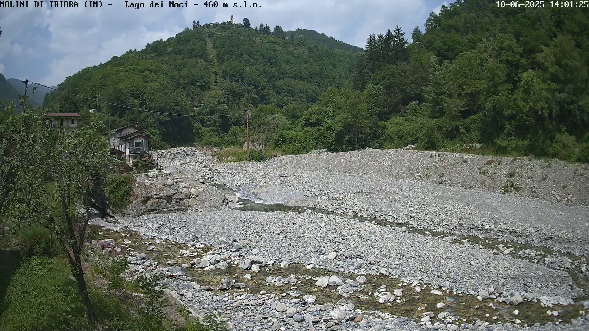 A mostly clear sky is over a rocky, dry riverbed with a small stream flowing through it, bordered by lush green vegetation and a couple of small buildings.