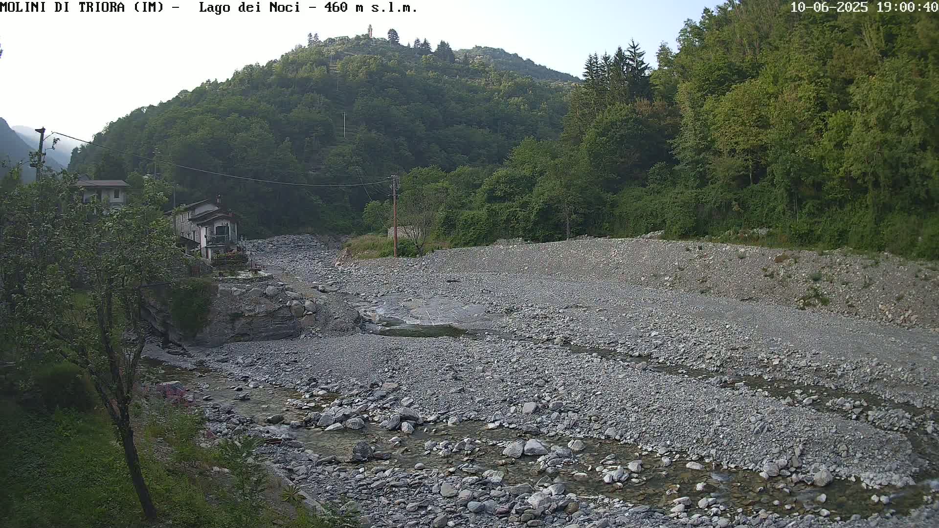 A mostly dry, rocky riverbed with a small stream flows through it, nestled between green hills under a mostly clear sky.
