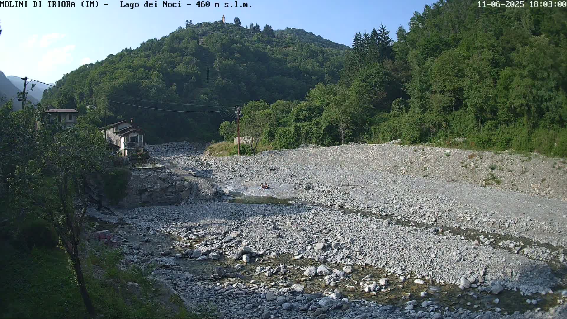 A rocky, dry riverbed with a small stream of water flows through it, near a few buildings and lush green hills under a clear sky.