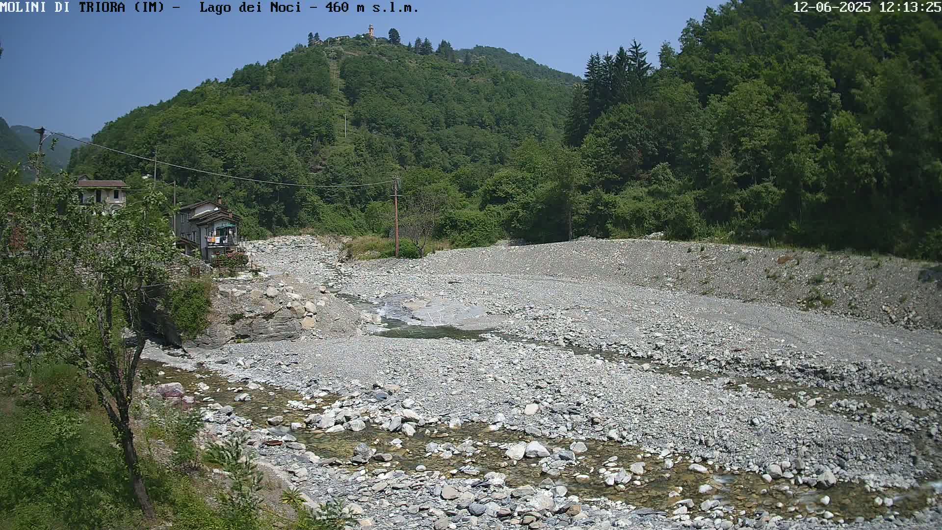 A shallow, rocky stream flows through a dry, gravelly riverbed, surrounded by lush green hills under a clear blue sky.