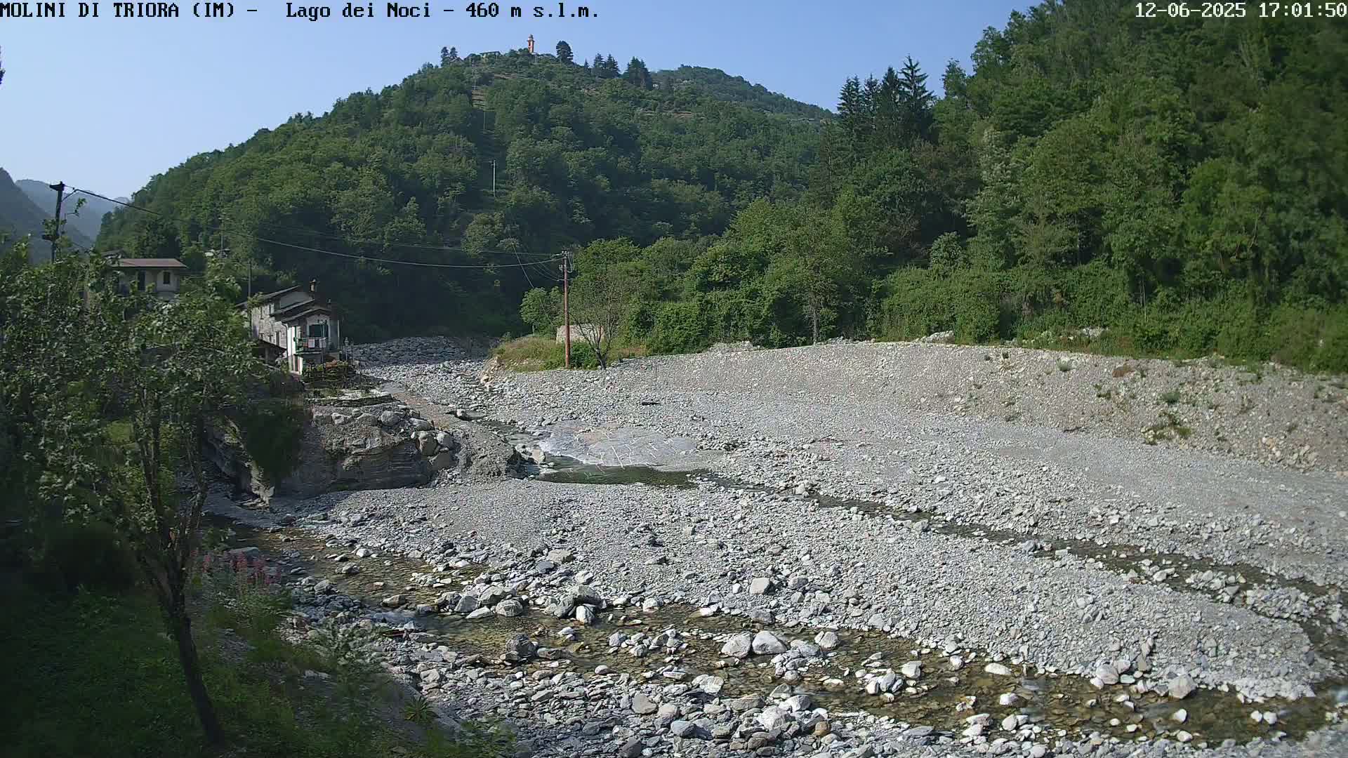 A shallow, rocky river flows through a dry riverbed beneath a clear sky and lush green hills.