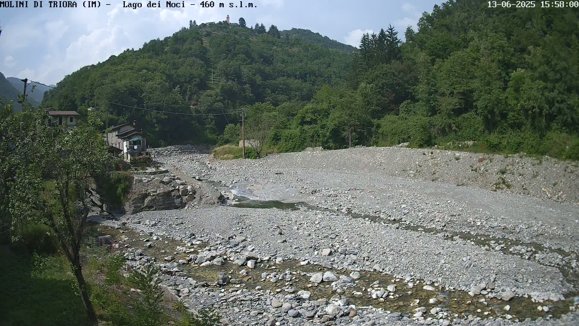 A mostly dry, rocky riverbed with a small stream running through it, nestled in a valley surrounded by lush green hills under a partly cloudy sky.