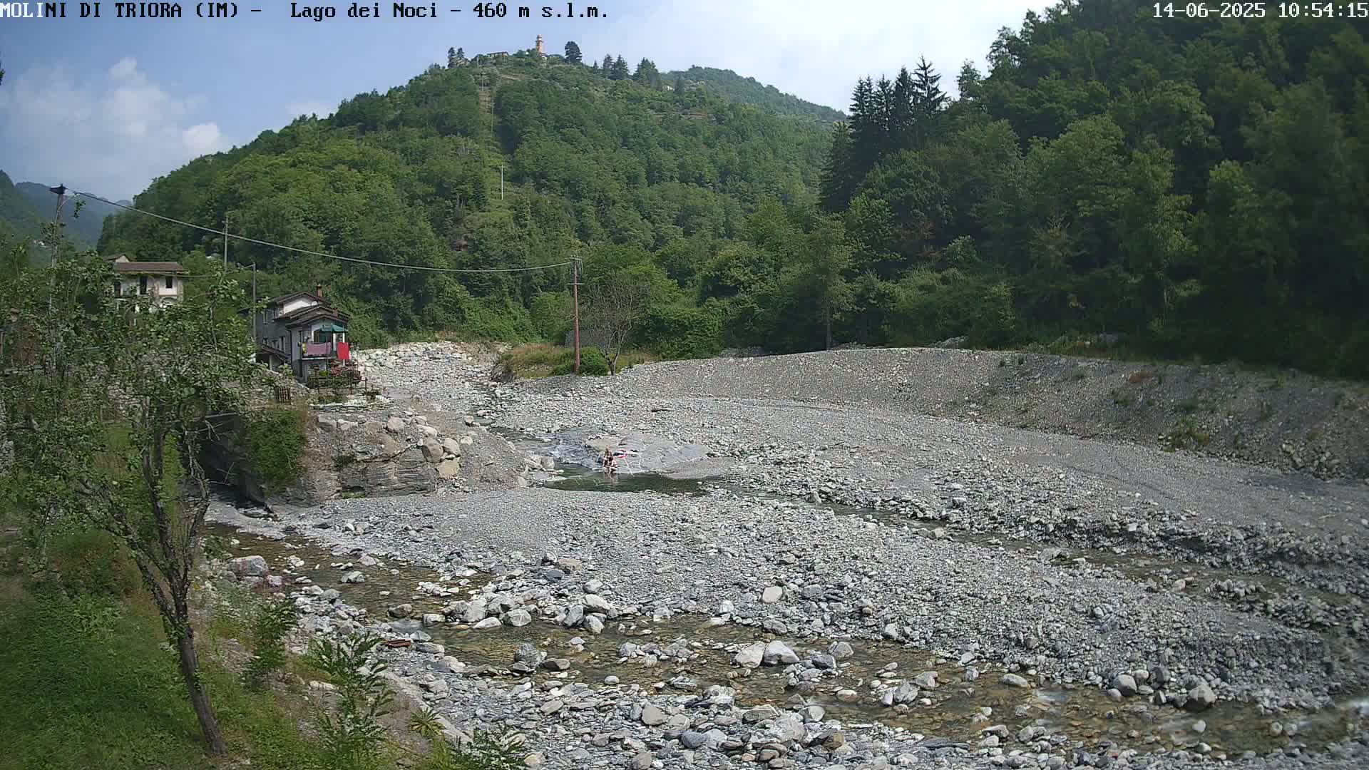 A rocky, mostly dry riverbed with a small stream of water flows through it, situated below a hillside covered in lush green trees under a partly cloudy sky.