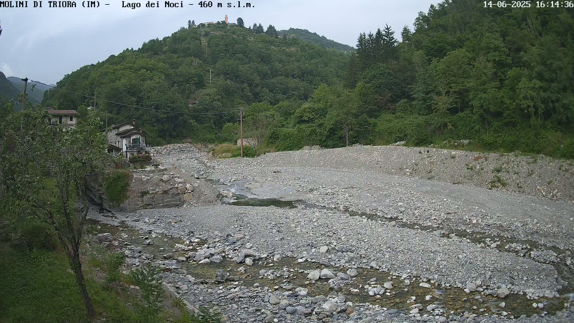A rocky, mostly dry riverbed with a small stream of water flows through it, nestled in a valley of lush green vegetation under an overcast sky.