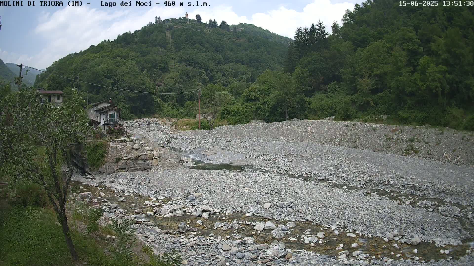 A mostly clear, partly cloudy day reveals a rocky, shallow riverbed with a small stream of water running through it, nestled within a valley surrounded by lush green hills and a small village.