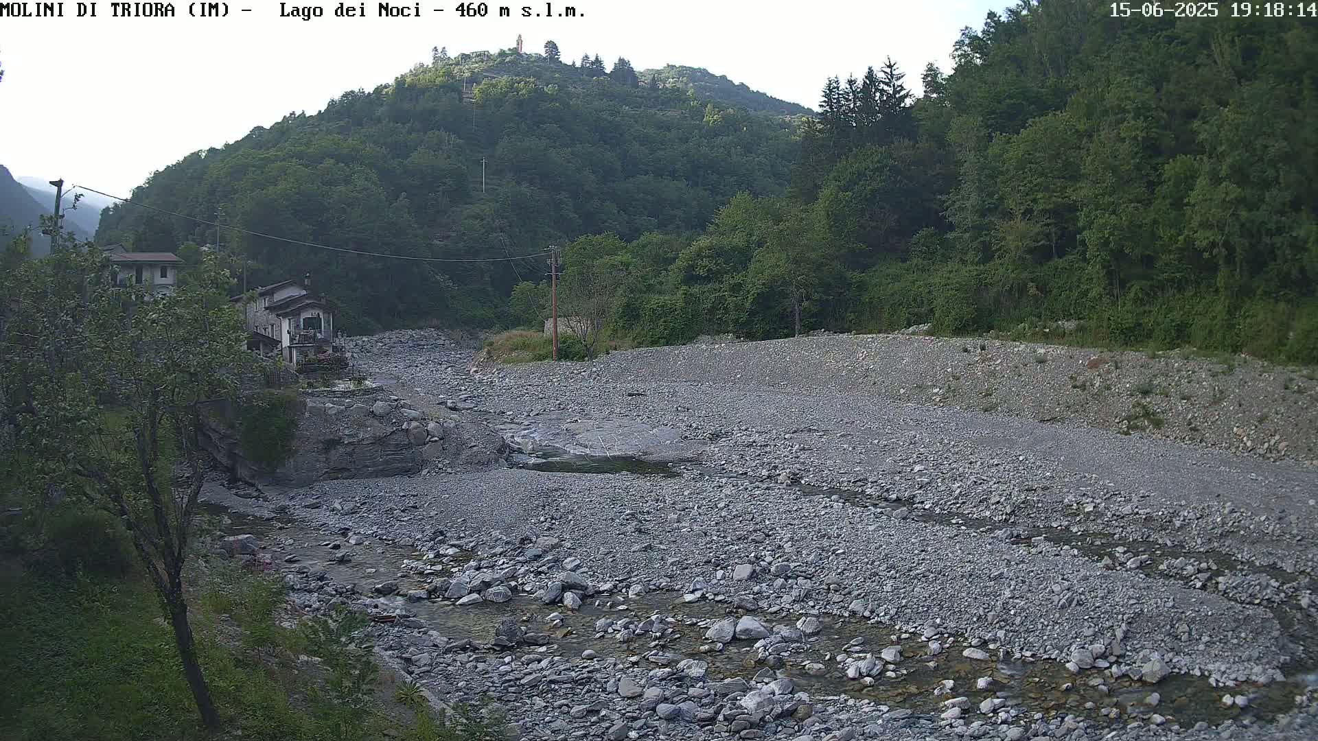 A rocky, mostly dry riverbed winds through a valley under a partly cloudy sky, past a small village nestled against a hillside covered in lush green trees.