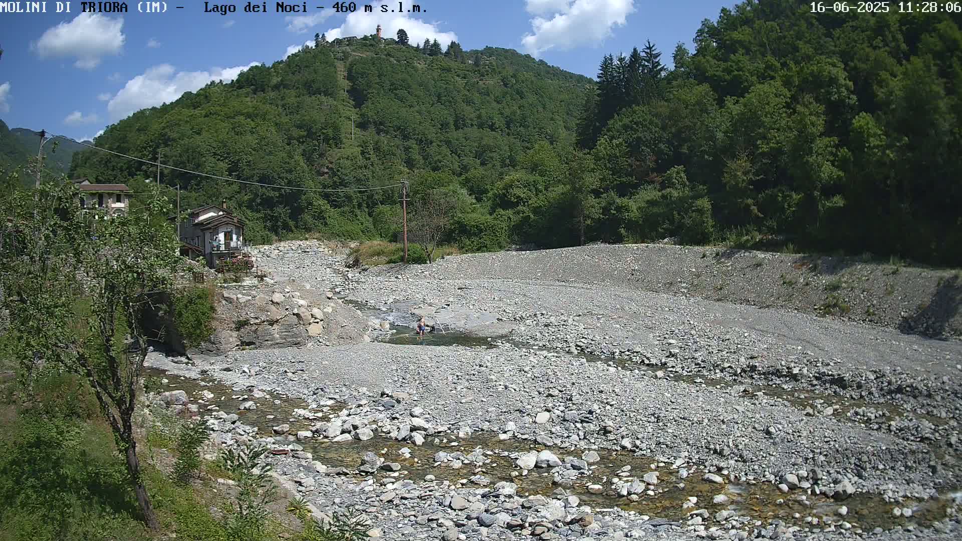 A rocky, mostly dry riverbed with a small stream of clear water flows through it, nestled in a valley surrounded by lush green hills under a partly cloudy blue sky.