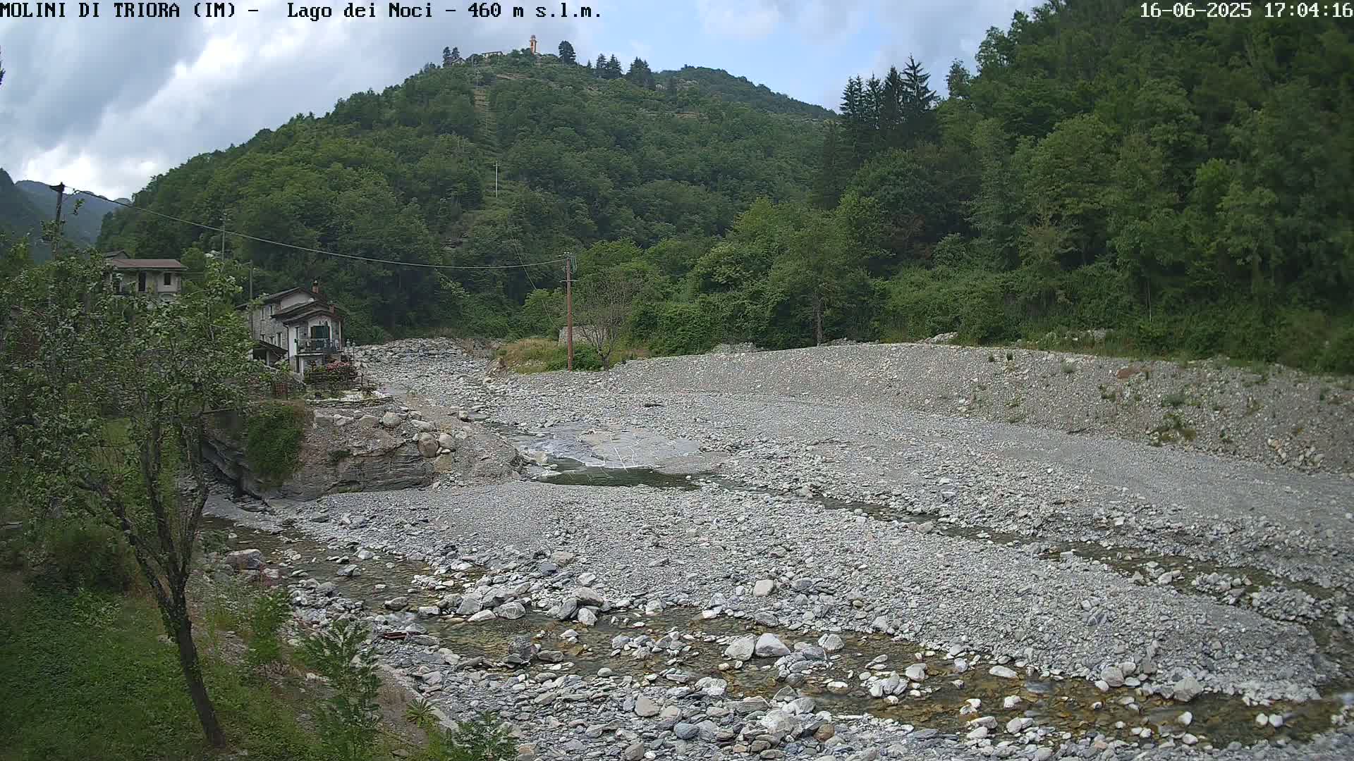 A mostly dry, rocky riverbed with a small stream flows through it, nestled in a valley surrounded by lush green hills under a partly cloudy sky.