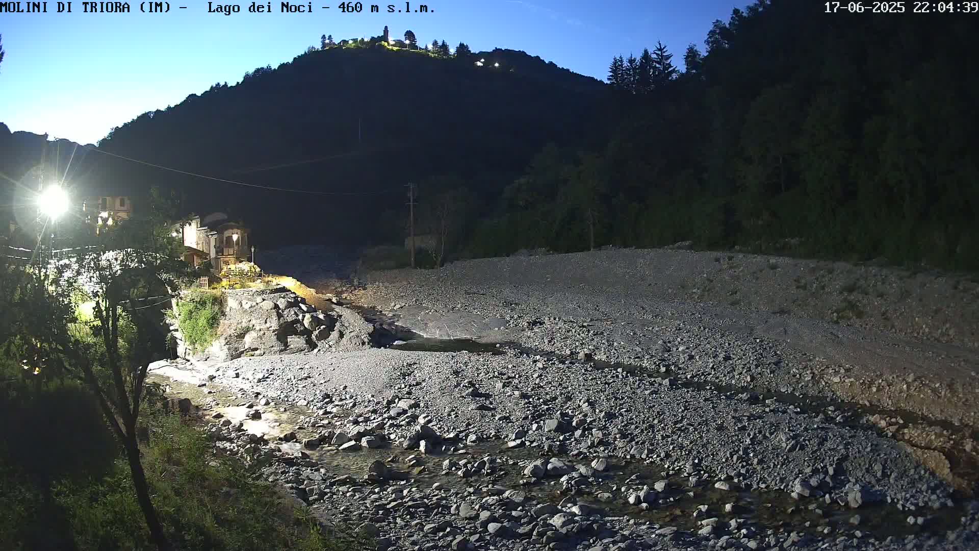 A rocky, shallow river flows through a dark, nighttime valley past a small village nestled against a hillside under a twilight sky.