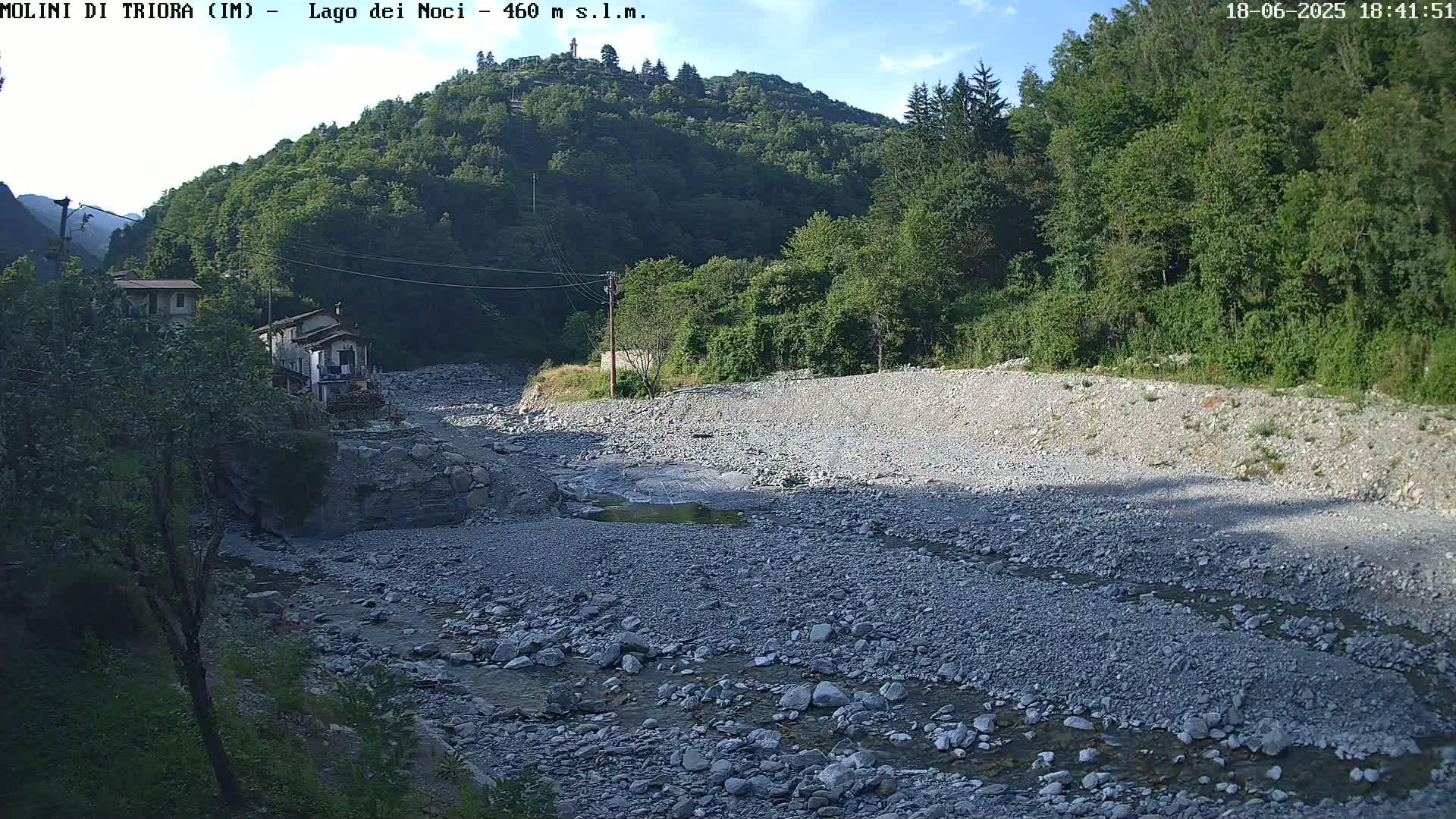 A mostly dry, rocky riverbed with a small stream flows through it, near a couple of houses nestled in a lush, green mountain valley under a partly sunny sky.