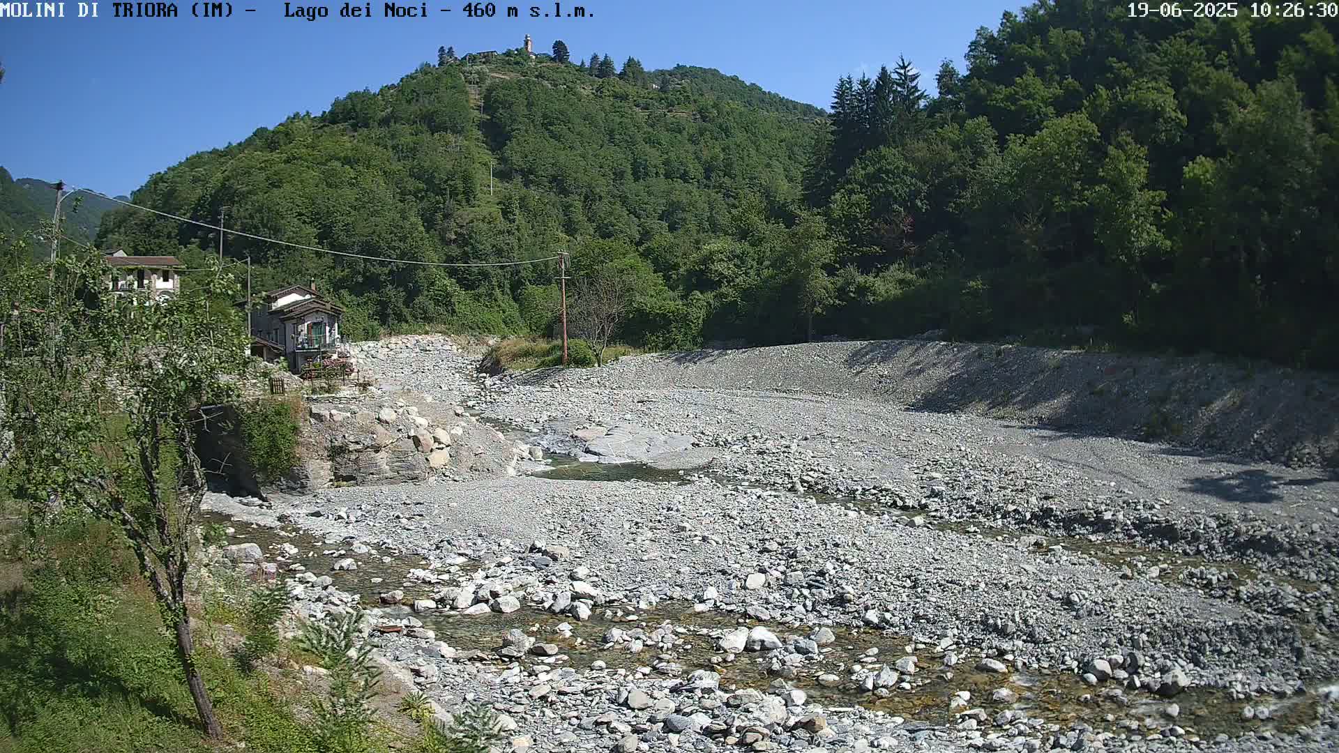 A rocky, dry riverbed with a small stream of water flows through it, nestled between green hills under a clear, sunny sky.