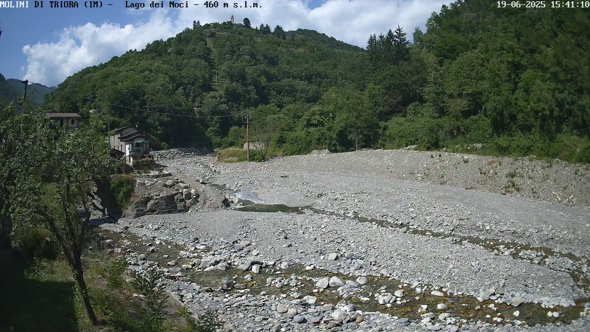 A mostly dry, rocky riverbed with a thin stream of water flows through it, nestled in a valley surrounded by lush green hills under a partly cloudy sky.