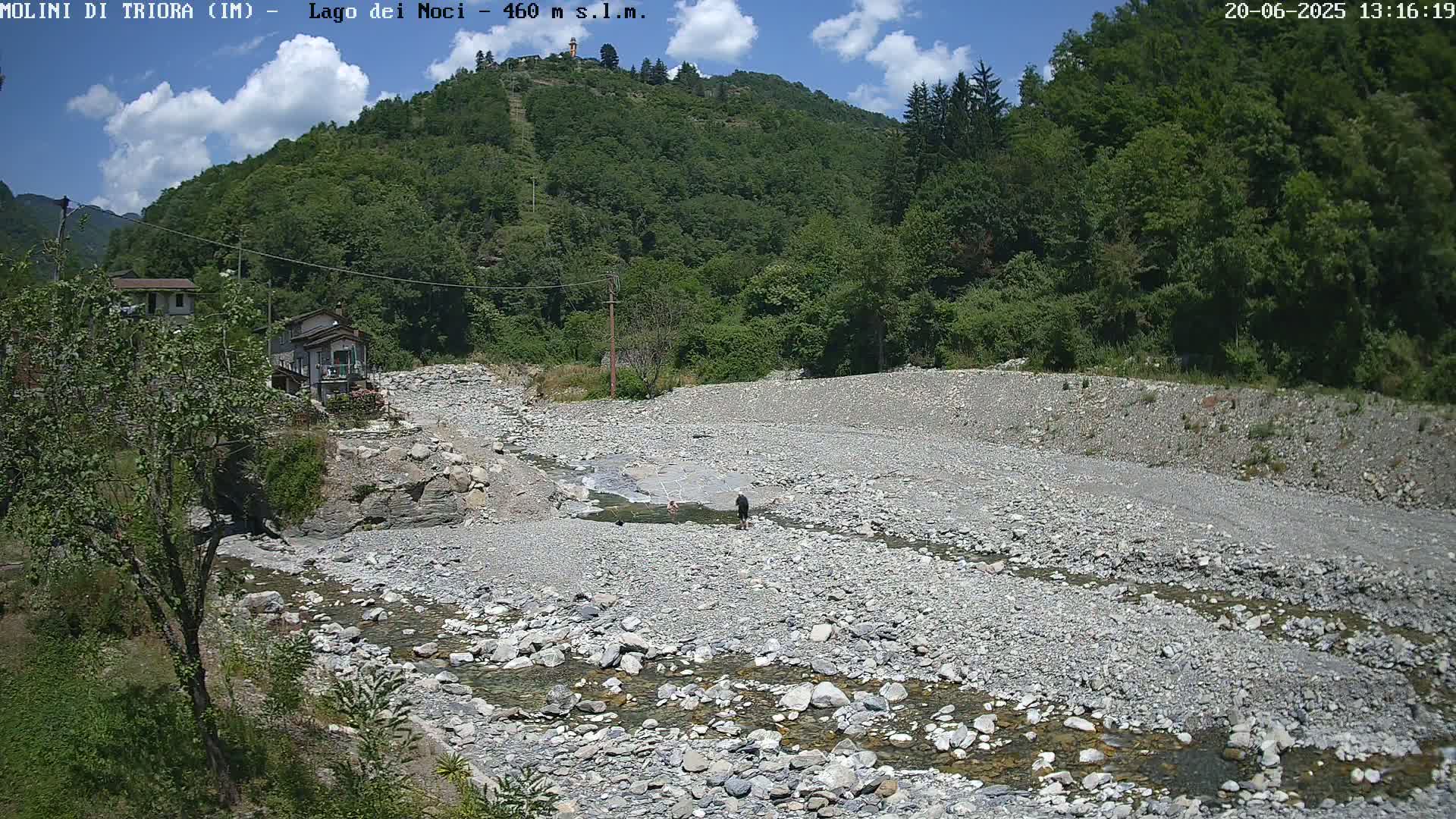 A shallow, rocky riverbed with a small stream of water flows through it, nestled between a hillside covered in lush green trees and a small village under a partly cloudy sky.
