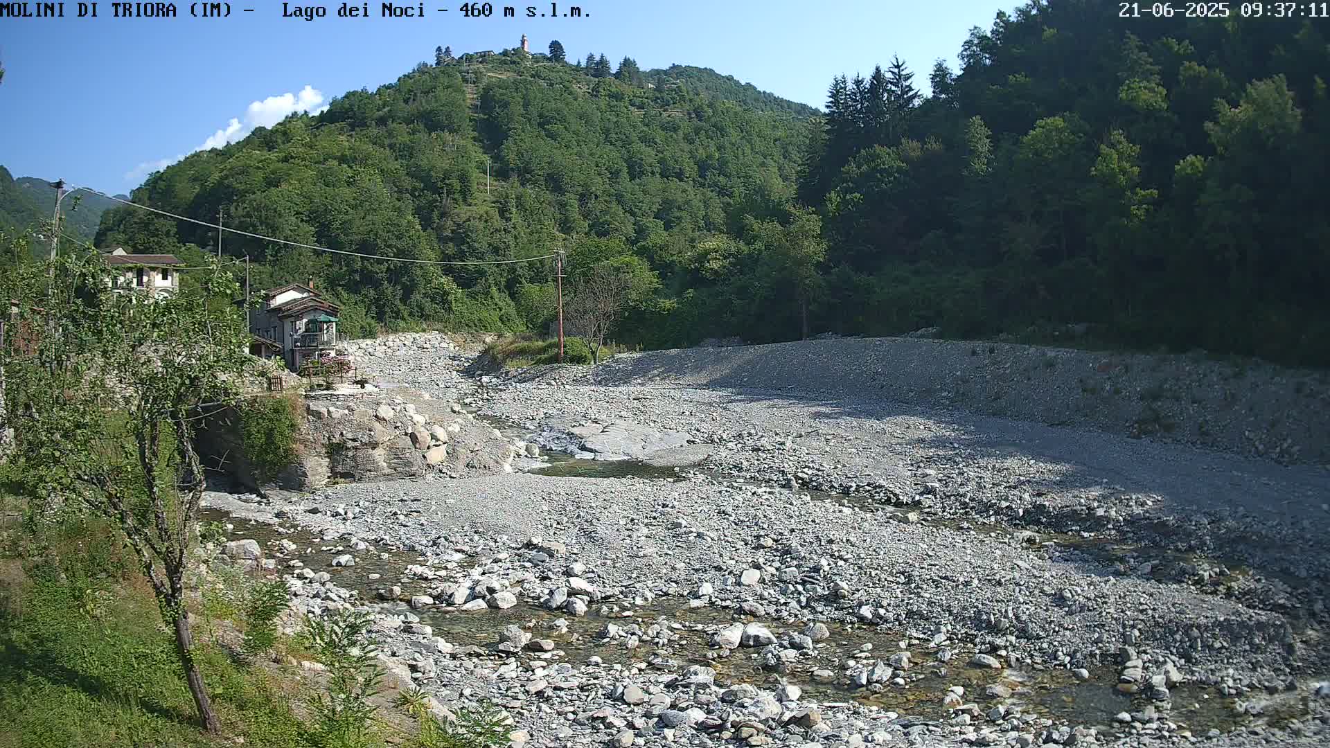 A mostly dry, rocky riverbed with a small stream of water flows through it, nestled between lush green hills under a clear blue sky.