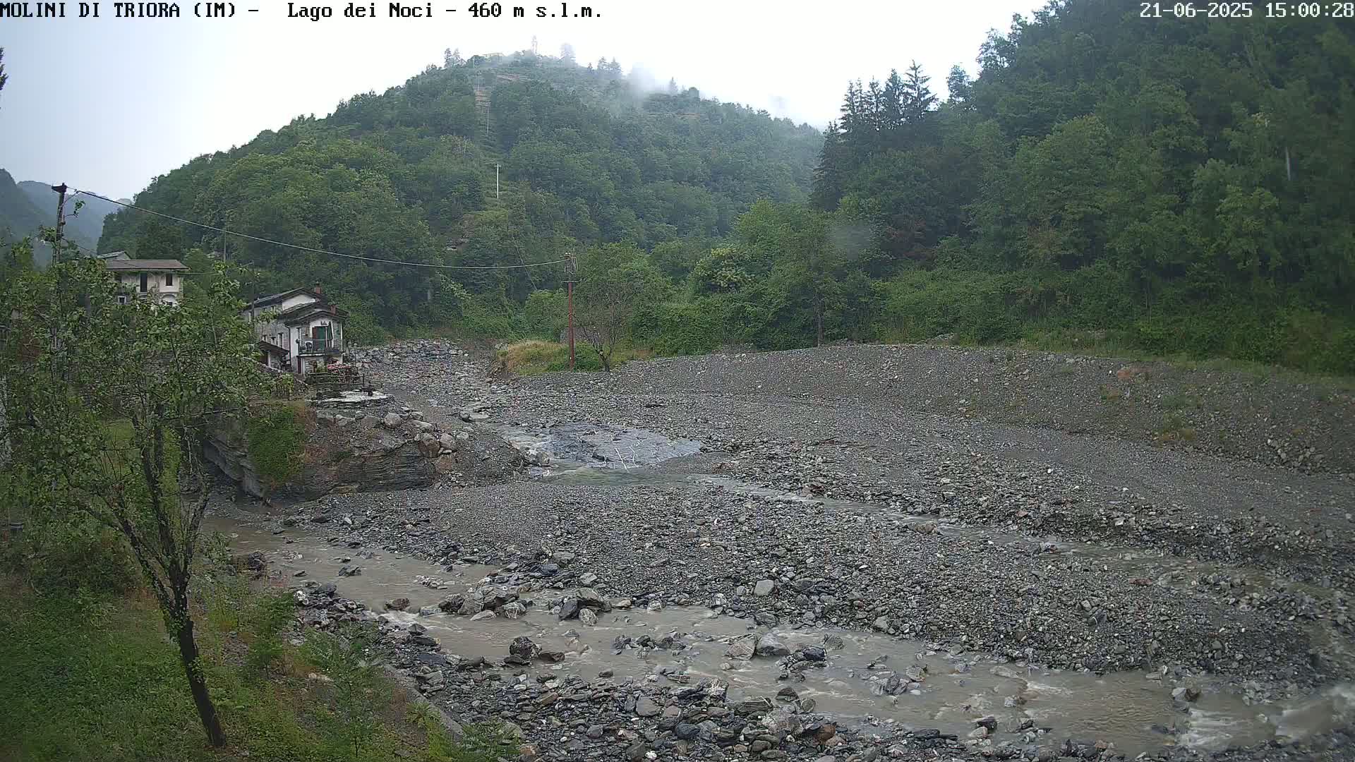 A rocky, shallow stream flows through a gravelly riverbed, bordered by lush green vegetation and buildings, under an overcast sky.