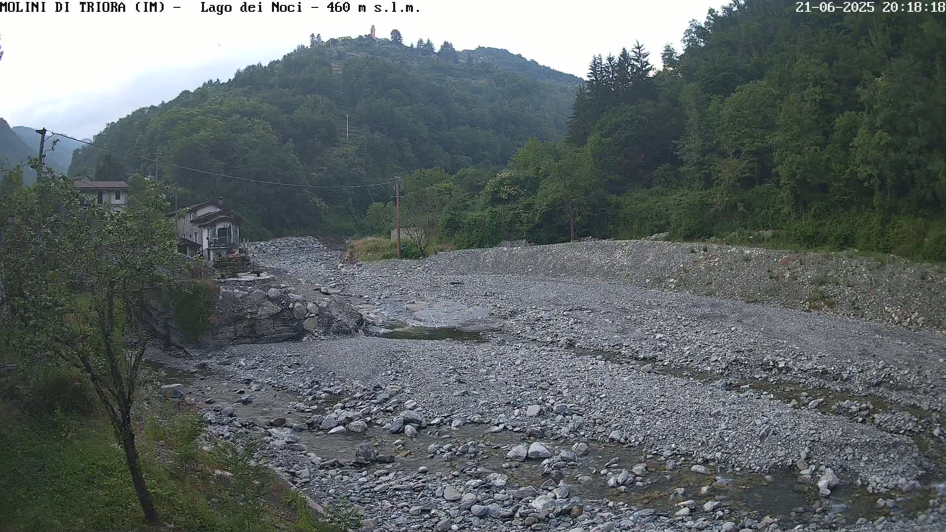A rocky, mostly dry riverbed with a small stream of water flows through it, nestled in a valley between lush green hills under a partly cloudy sky.