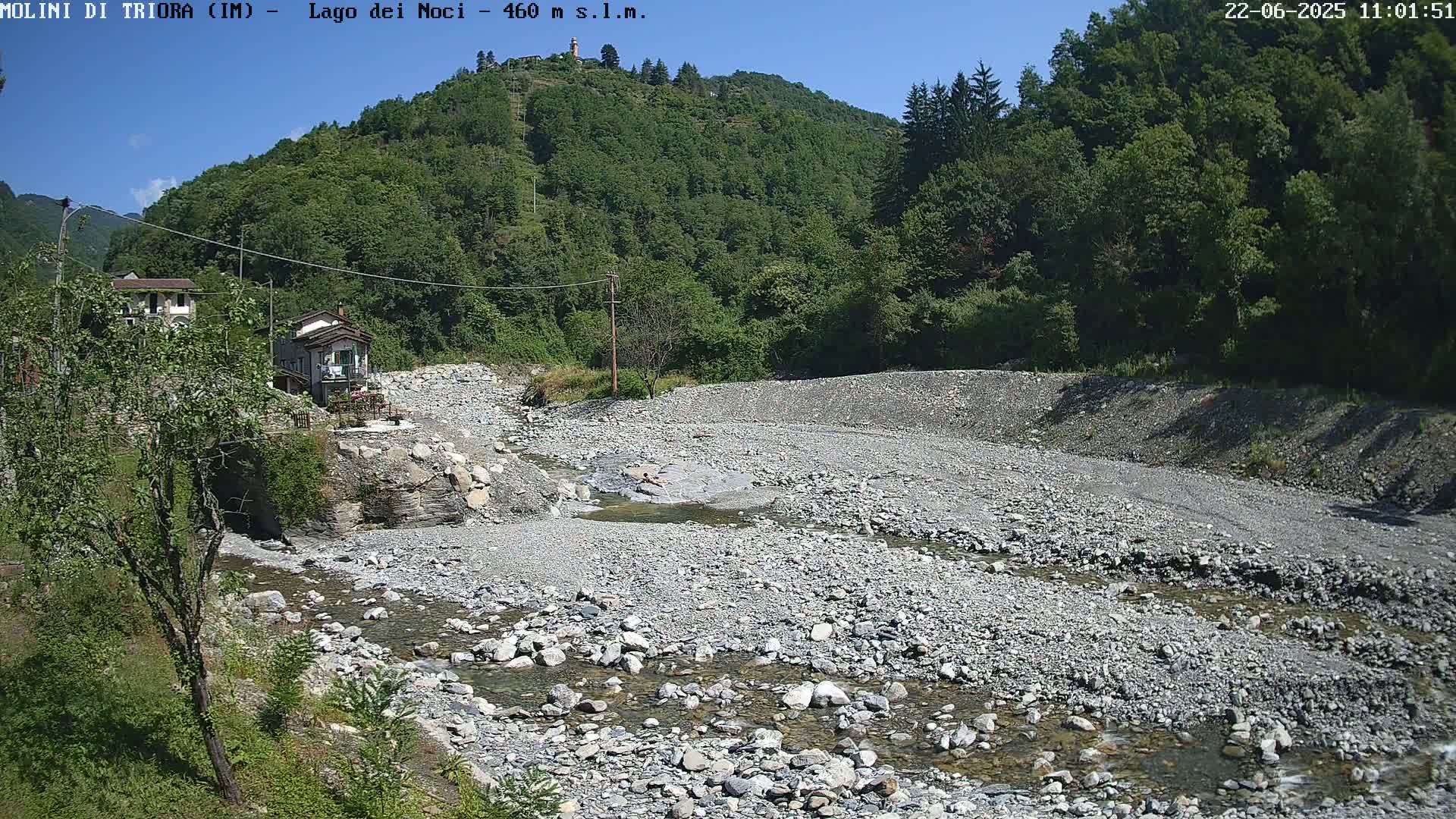 A rocky, mostly dry riverbed with a small stream flows through it, nestled in a valley between lush green hills under a clear blue sky.