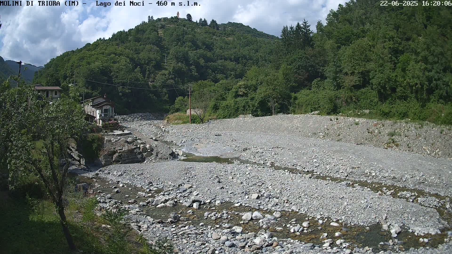 A mostly dry riverbed filled with rocks and a small stream of water runs through a valley under a partly cloudy sky, with green hills and a few buildings visible in the distance.
