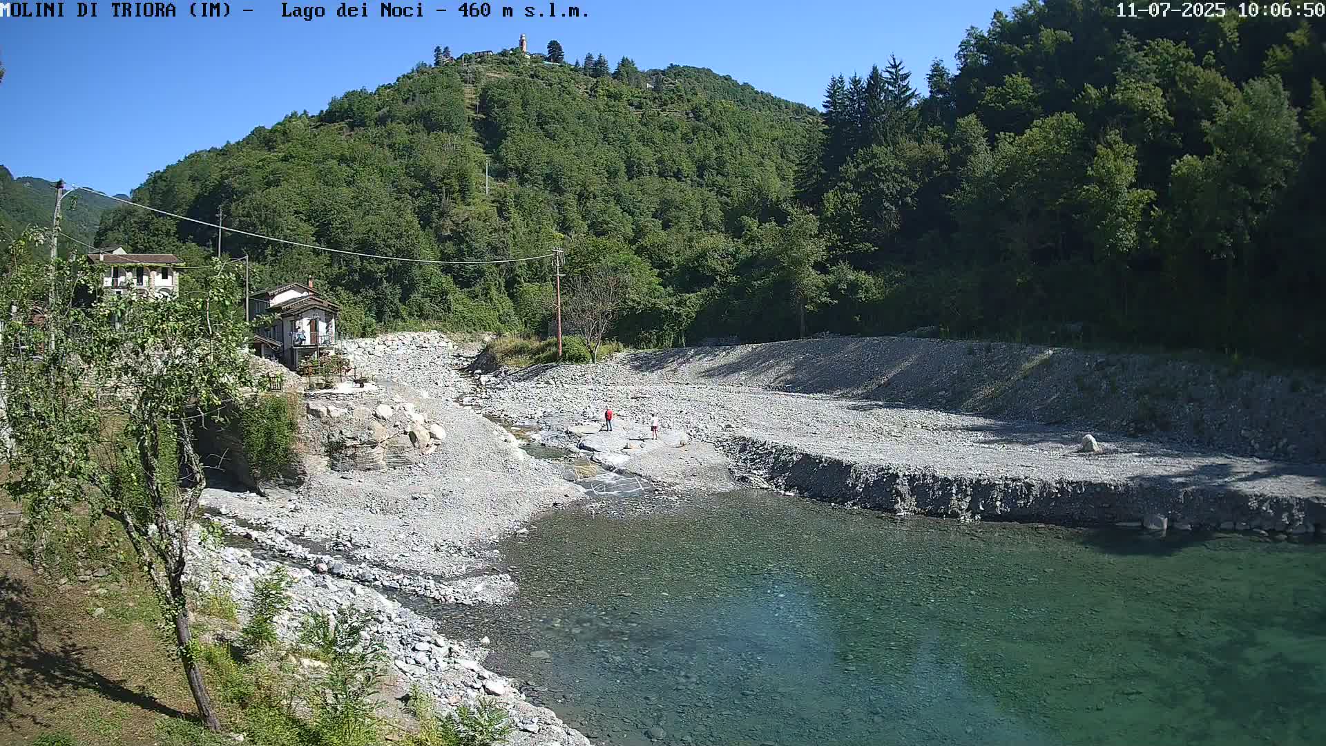 A shallow, clear river flows through a rocky, dry riverbed surrounded by lush green hills under a bright, sunny sky.