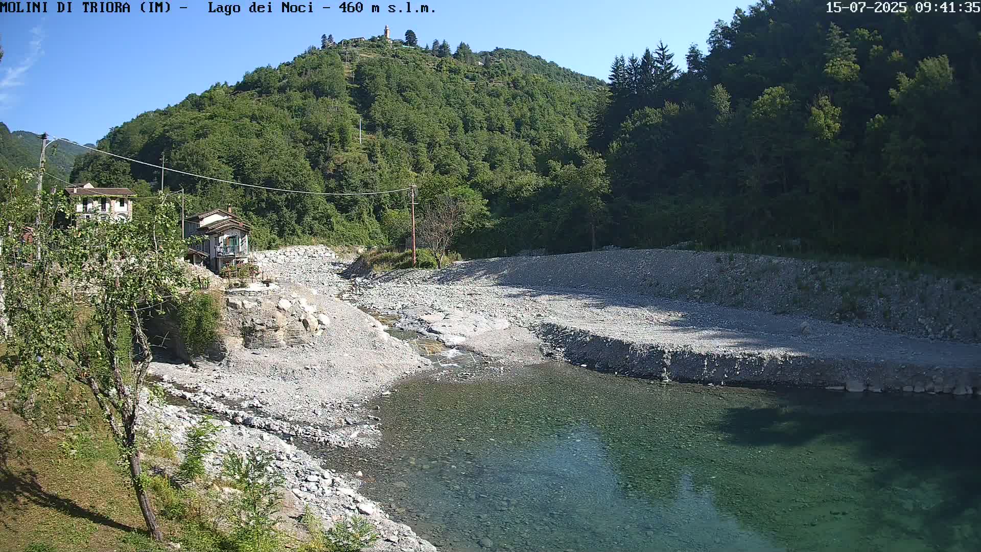 A shallow, clear river flows through a rocky riverbed, bordered by lush green hills under a clear, sunny sky.