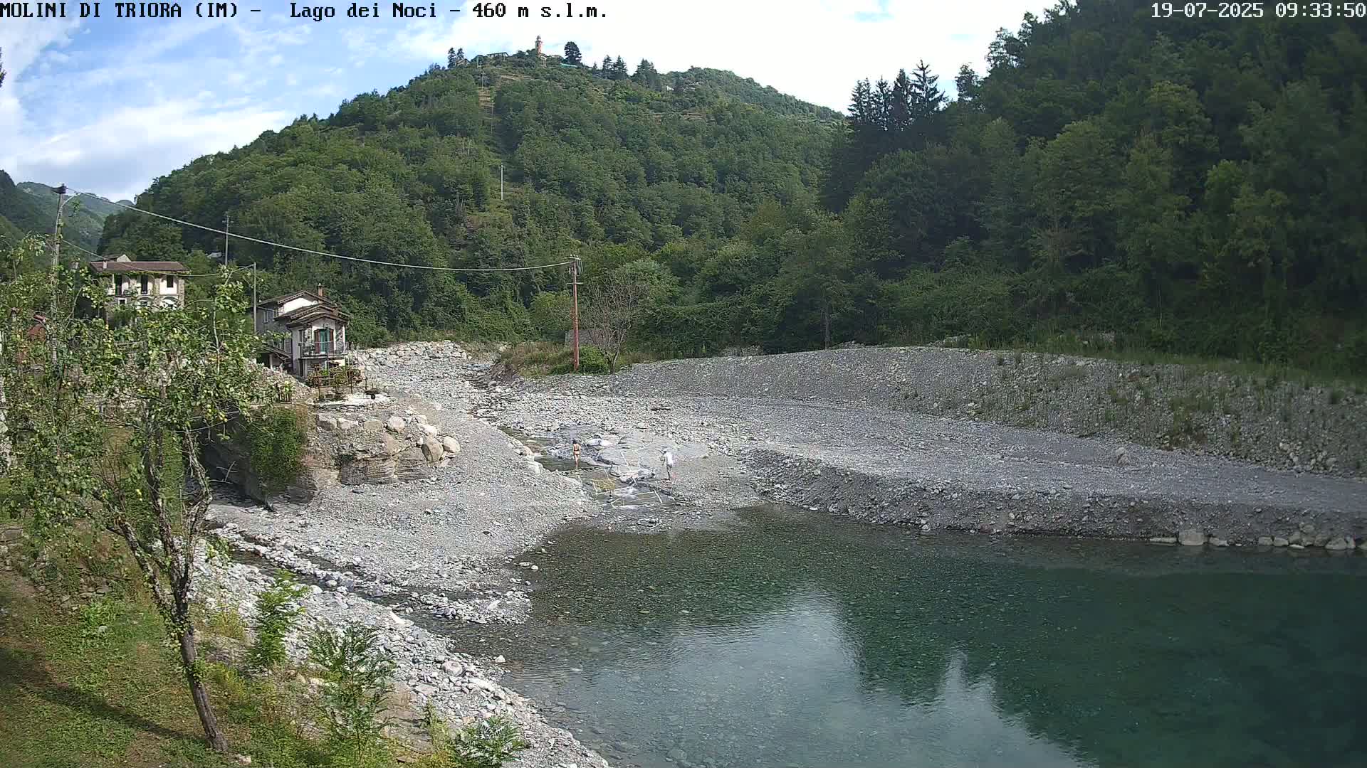 A rocky riverbed with a calm, teal pool of water flows beneath a hillside covered in lush green trees under a partly cloudy sky.