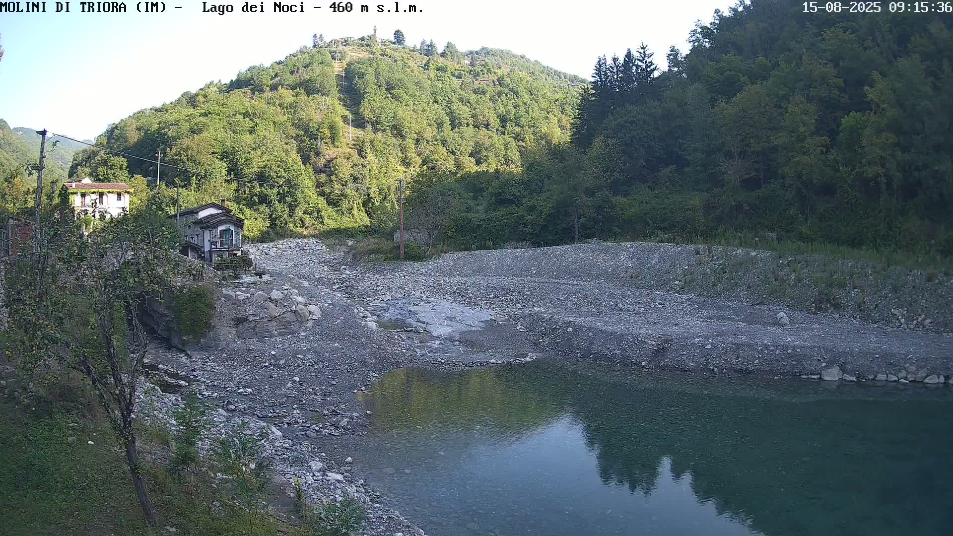 A calm body of water reflects the surrounding hillsides, which are covered in lush green vegetation under a clear sky, with a rocky, dry riverbed in the foreground.