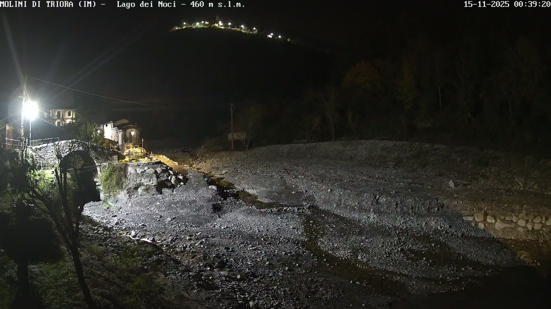 A nighttime scene depicts a historic stone bridge spanning a wide, rocky riverbed with a small stream, bordered by illuminated buildings and distant lights on a dark hillside, all under clear conditions.