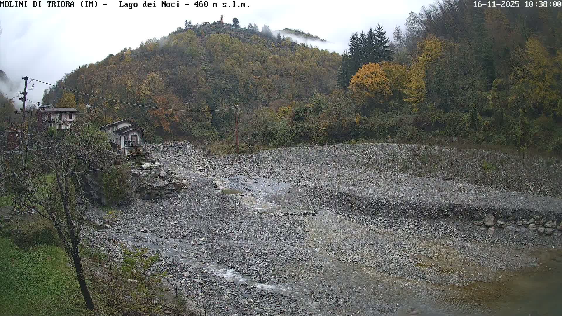 On a cloudy and misty autumn day, a rocky riverbed with a shallow stream flows through a valley lined with houses and hillsides covered in trees displaying fall colors.