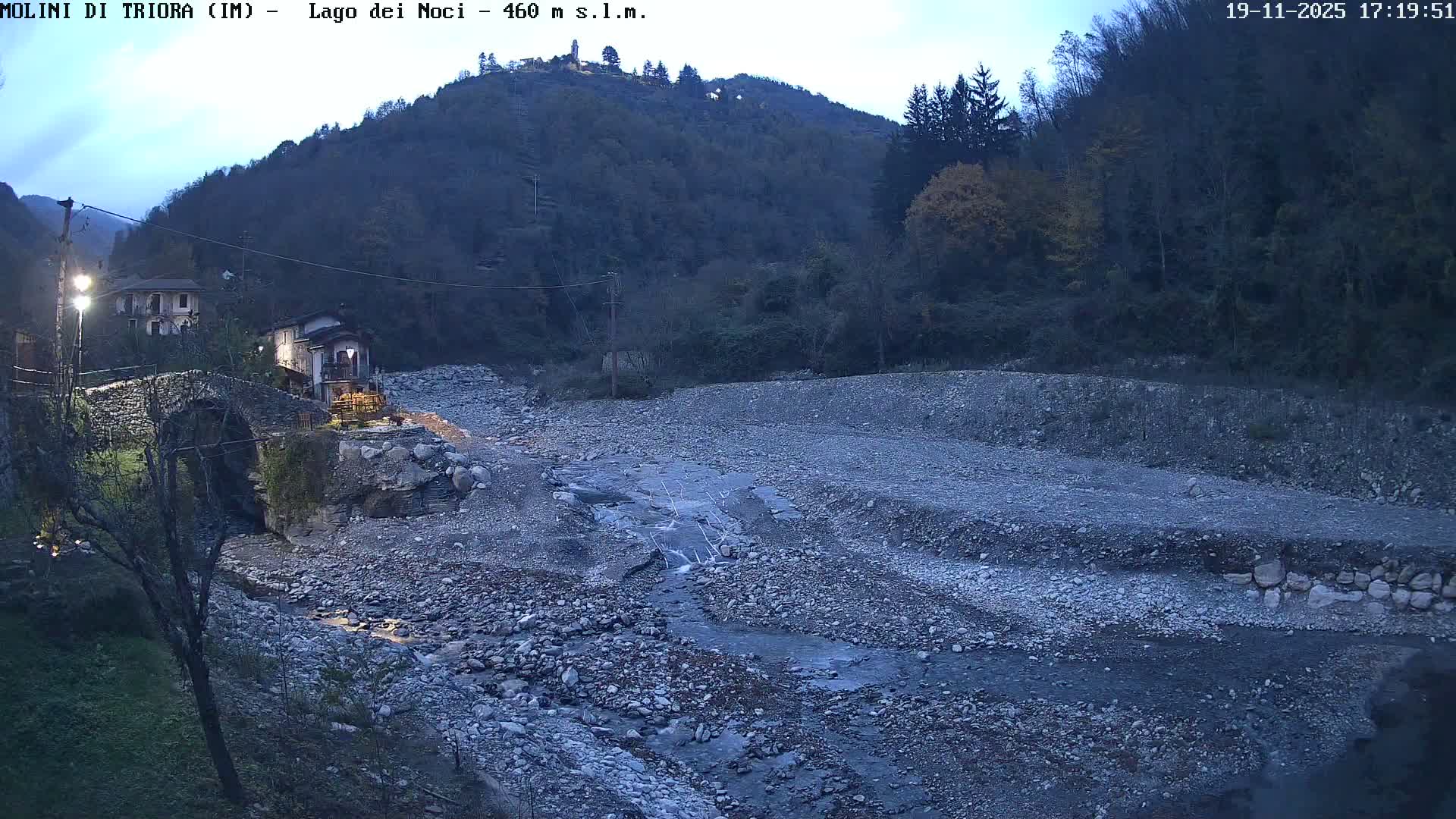 On a cloudy and misty autumn day, a rocky riverbed with a shallow stream flows through a valley lined with houses and hillsides covered in trees displaying fall colors.