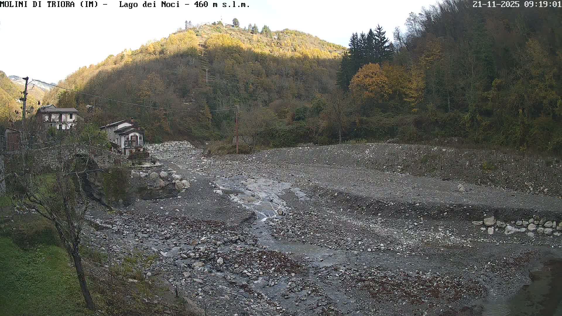 A narrow stream winds through a wide, rocky riverbed in a valley, flanked by old stone buildings on the left and forested hills displaying autumn colors, all under clear daylight.