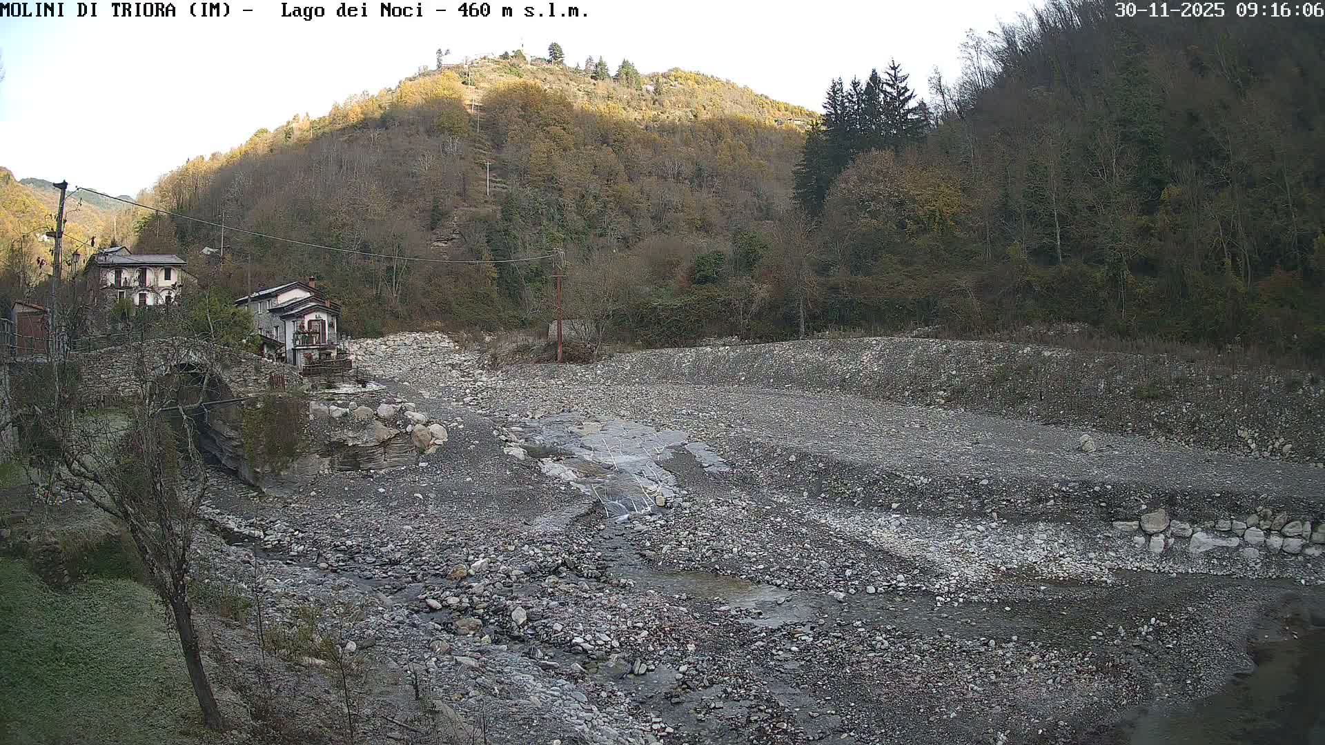 A wide, rocky riverbed with a small stream flows through a valley, flanked by old buildings and a stone bridge on the left and hillsides covered in mostly bare trees, under bright, cool daylight.
