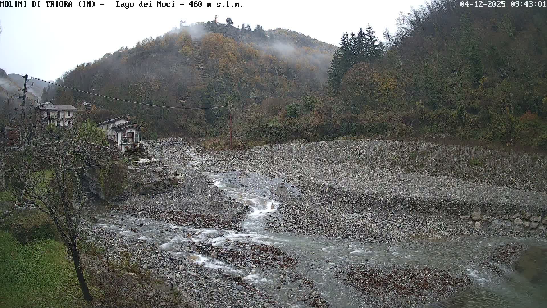 A fast-flowing river winds through a rocky valley floor on a misty, overcast day, flanked by wooded hillsides with sparse autumn foliage and a few buildings visible on the left.